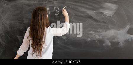 Rear view of a student or teacher with long brunette hair writing on a blank chalk board or blackboard with copyspace English alphabet. Back to school Stock Photo