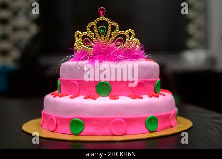 Two-tier fondant cake design with a tiara on top, decorated with icing flowers. Delicious pink-red color-themed round cake on yellow cake tray against Stock Photo