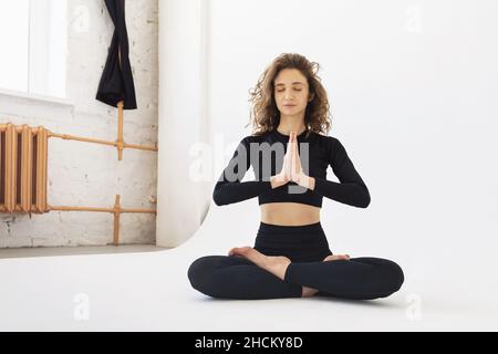 A young beautiful woman practicing yoga, performs meditation, sits in a lotus pose with namaste, trains in black sportswear in a light room Stock Photo