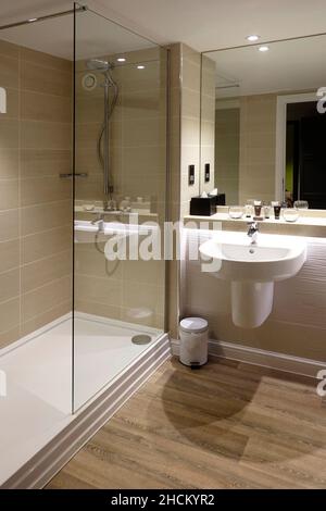 Modern walk in tiled shower & screen large safety grab rail in en suite hotel bathroom full height mirror over wall mounted wash hand basin England UK Stock Photo