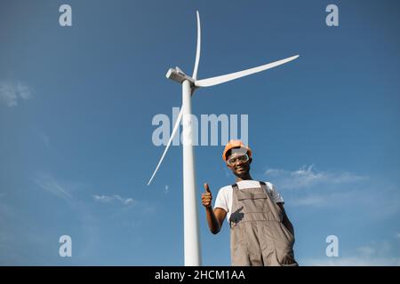 African american engineer in uniform and helmet gesturing with fingers thumbs up with while standing on eco farm. Huge wind turbine on background. Production of green energy. Stock Photo