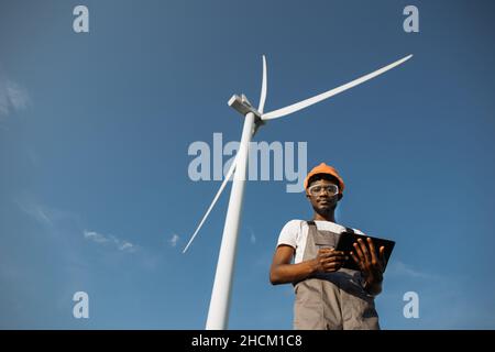 Competent technician using digital tablet while controlling working process of wind turbines outdoors. African american man wearing grey overalls, orange helmet and safety glasses. Stock Photo