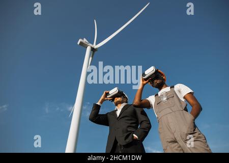 Indian businessman in suit and african american engineer in uniform using VR goggles for work on farm with wind turbines. Production of green energy and modern technology concept. Stock Photo
