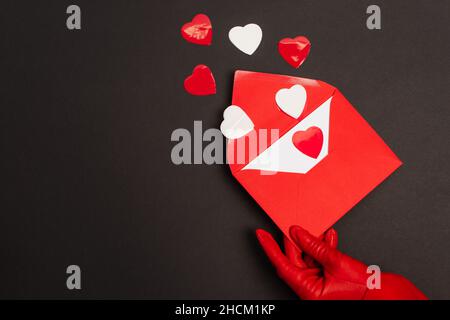 cropped view of person in red glove holding envelope with love letter and paper cut hearts on black Stock Photo