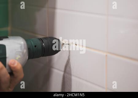Close-up of man hands with drill making holes for dowels into tiled wall in washroom causing loud noise. Renovation works in bathroom. Dirt from Stock Photo