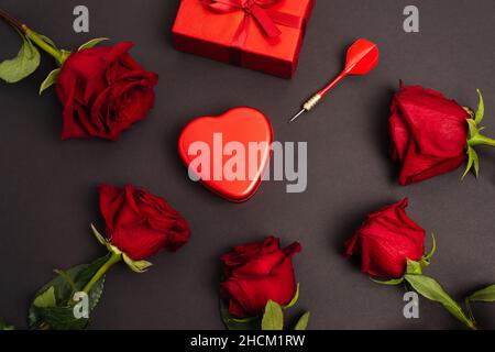 flat lay of metallic heart-shaped box near wrapped present, dark and roses, on black Stock Photo