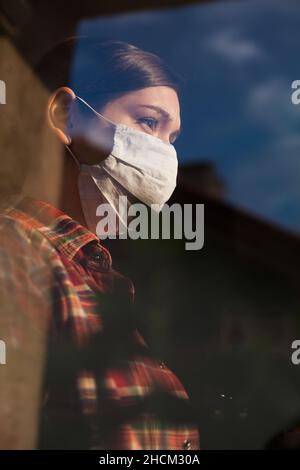 Profile portrait of young caucasian female wearing protective face mask,standing by the room window looking outside,Coronavirus pandemic lockdown Stock Photo