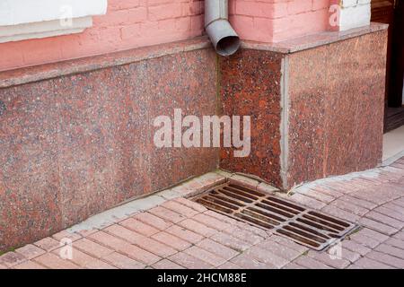 drainage of rainwater a pipe on the wall of the facade of the building with a rain grate of the hatch on the pedestrian sidewalk made of stone tiles n Stock Photo