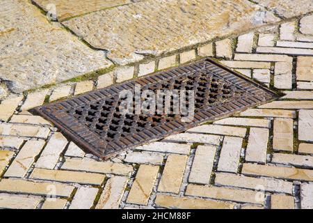 iron rusty rectangular manhole with shaped holes in form of crosses in lid on the sidewalk paved with brick tiles, retro sewer hatch on a stone sidewa Stock Photo