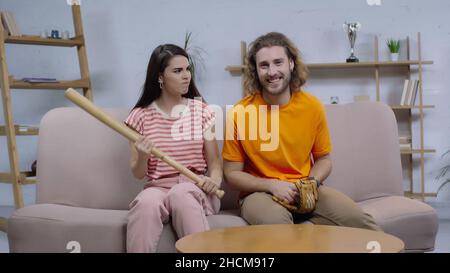 angry woman with baseball bat looking at happy man watching match on tv at home Stock Photo