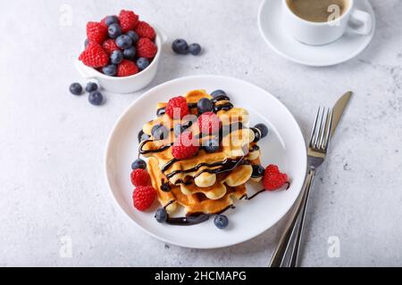 Viennese or Belgian waffles with fresh berries (raspberries and blueberries) and chocolate sauce on a white plate and a cup of coffee. Traditional des Stock Photo