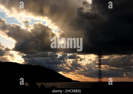 Dark storm clouds at sunset, dark storm clouds with sunrays, mountain and radio tower silhouette over the sea. Climate and storm concept Stock Photo