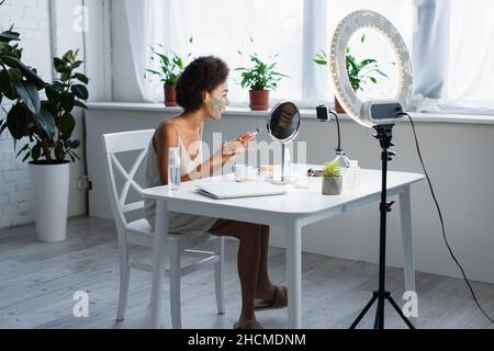 Side view of african american blogger with clay mask on face holding cosmetic brush near smartphone Stock Photo
