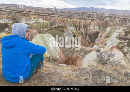 Person with hood and blue jacket sits and calmly wit jot looking around the the landscape of red valley in Cappadocia. Stock Photo