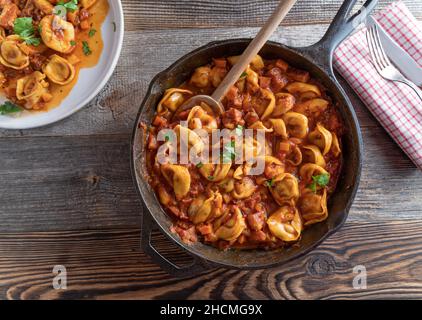 Italian pasta dish with tortellini in a delicious tomatoe, vegetable bacon sauce. Served in rustic pan isolated on wooden table background. Overhead Stock Photo
