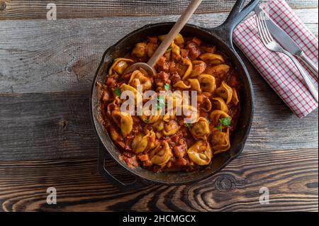 Italian pasta dish with tortellini in a delicious tomatoe, vegetable bacon sauce. Served in rustic pan isolated on wooden table background. Overhead Stock Photo