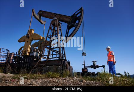 Against cloudless sky operator standing near oil pipeline with spanner and preparing to servicing petroleum borehole. Oilman in overalls monitoring serviceability of drilling pump rig. Stock Photo