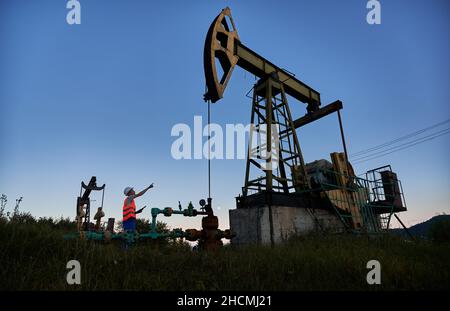 Side view of capable inspector pointing at pump derrick jack while checking serviceability at oil station, professional male worker in uniform extract natural resources using drilling system Stock Photo