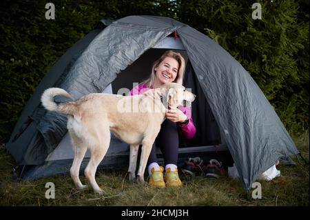 Outdoors camping. Young happy woman with smile on her face sitting in gray tourist tent and caressing her dog. Active vacation of female hiker with pet in nature. Stock Photo