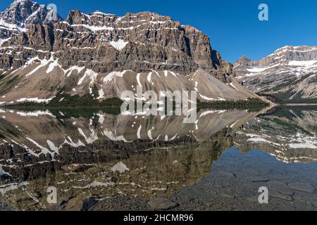 The sheer rock wall of Crowfoot Mountain with a dusting of snow reflected by Bow Lake along the Icefields Parkway at Banff National Park in Alberta, Canada. Bow Lake is formed from the glacier melt of the Crowfoot Glacier and Wapta Icefields. Stock Photo