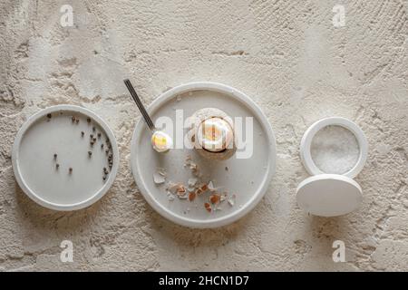 One boiled egg in stoneware egg cup with egg shell broken pieces in concrete tray, salt flakes and pepper in white concrete bowls on rough textured cl Stock Photo