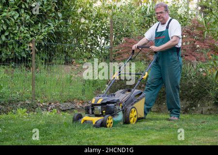 Man pushing lawnmower through small backyard in spring. Front view of man with overall. Stock Photo
