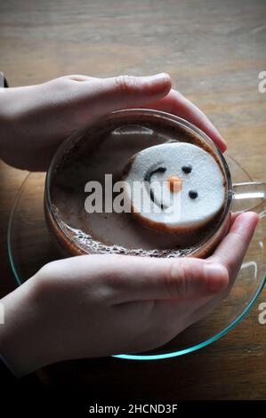 A child's hand cradling a hot chocolate in a glass mug with saucer and a snowman face marshmallow Stock Photo