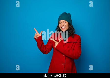 Charming smiling curly dark-haired woman in bright red coat and woolen green hat poses against blue background, looks at camera and points with finger Stock Photo