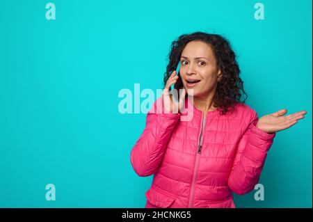 Surprised shocked emotional curly haired beautiful woman in bright pink jacket talks on mobile phone, and gestures with her hand palm up, isolated on Stock Photo