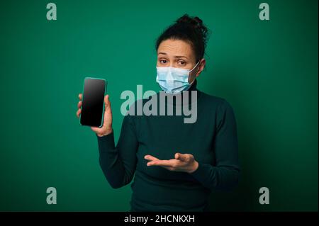Portrait of a serious millennial woman in protective face cover looks at camera and shows her smartphone with empty blank screen for copy space, isola Stock Photo