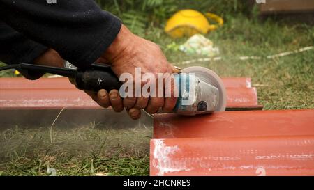 Roofer using an angle grinder machine to cut a roof tile of red color. Close up of male worker hands cutting a roof tile with a professional saw. Stock Photo