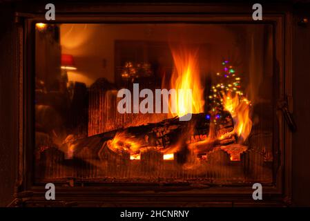 Wood logs burning in a fireplace for Christmas. A Christmas tree decorated with light tinsels is reflected on the glass of the fireplace. Stock Photo