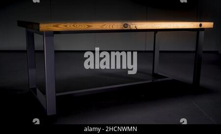 Wooden table with steel legs on dark grey background. Abstract table with brown wooden polished surface standing in the room where the light turns off Stock Photo