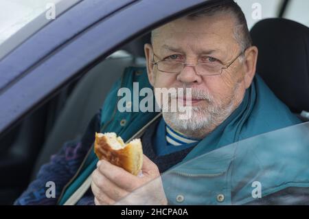 Bearded Caucasian senior driver taking patty and looking in camera while sitting inside his car Stock Photo