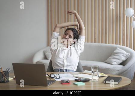 Tired italian lady remote employee stretching arms while sitting at desk with laptop Stock Photo