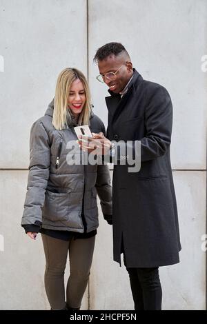 Couple smiling and looking at cell phone near a gray wall of a building. Boy showing his girlfriend his phone screen. Stock Photo