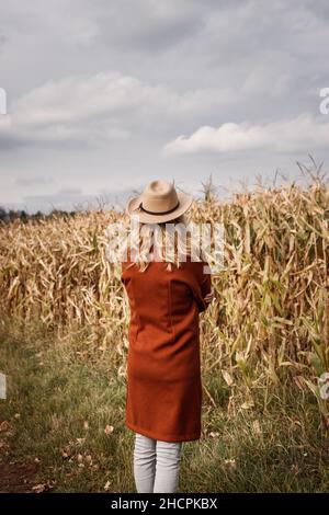 Stylish woman wearing red coat and hat is standing next corn field. Trendy autumn fashion collection. Elegance at fall season outdoors. Vintage pastel Stock Photo