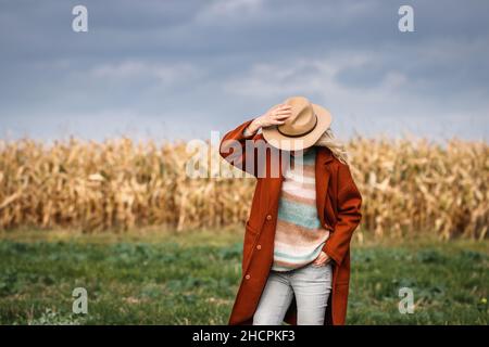 Trendy autumn fashion collection. Stylish woman wearing red coat, sweater and hat is standing next corn field. Elegance at fall season outdoors. Stock Photo