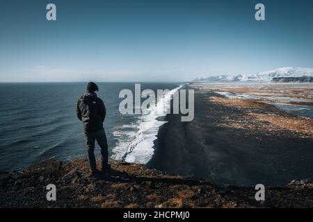 Person standing at Dyrholaey lighthouse in Iceland looking out over the black sand beach below during winter with snow and beautiful sunny weather. Stock Photo
