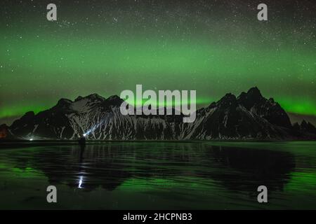 Young traveler standin in front of Vestrahorn Stockknes mountain range with aurora borealis and reflection at the beach in Iceland. One of the most Stock Photo