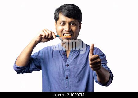 A young Indian male in blue dress showing thumbs up while brushing teeth on white background Stock Photo