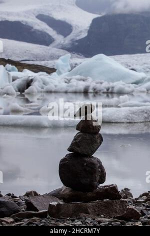 Small man-made artistic tower of smooth rocks balanced on top of each other beside a melt water stream in Iceland with ice and snow in the background Stock Photo