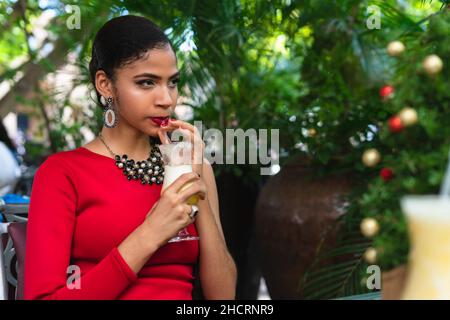 young latin woman in elegant red dress sitting outside drinking a refreshing drink Stock Photo