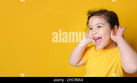 Little Asian girl holds her hand near her ear and listenings. Exciting face on Asian child girl. Exciting face on happy asian girl wear yellow shirt a Stock Photo