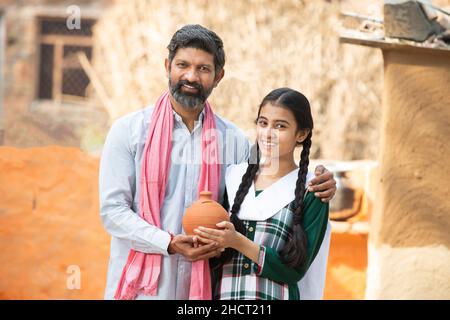 Portrait of happy Indian farmer father and daughter holding clay money box or gullak, Smiling beard man and young girl traditional piggy bank, dad tea Stock Photo