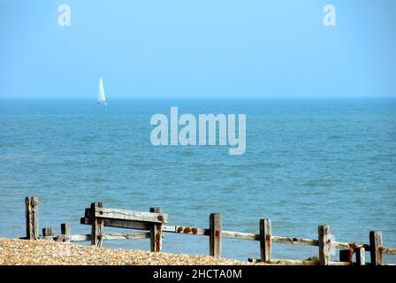 Lone yacht with white sail, Bermuda rig, sailing in the English Channel off Pevensey Bay, East Sussex, England and old wooden breakwater in foreground Stock Photo