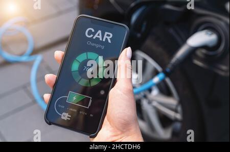 Woman Charging Electric car with Cable Looking at App On Mobile Phone. Close up of smartphone screen. Hand holding smart device. Mobile application fo Stock Photo