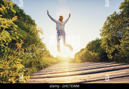 Happy young man jumping with raised hands and legs, and enjoying life over a Wooden jetty at sunset Stock Photo
