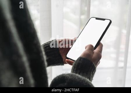 Female hand holding black cellphone with white screen at home against a window, copyspace for your individual text. Stock Photo