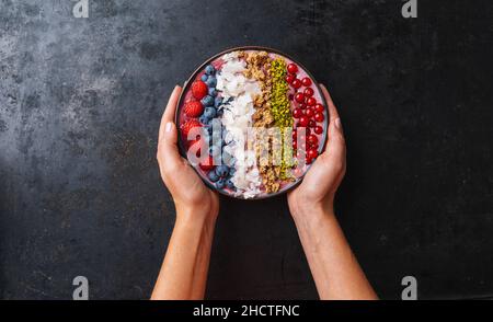 Woman hands hold a Healthy breakfast bowl, wirh blueberry smoothie with, raspberry, blueberrys, coconut, nuts and currants toppings Stock Photo